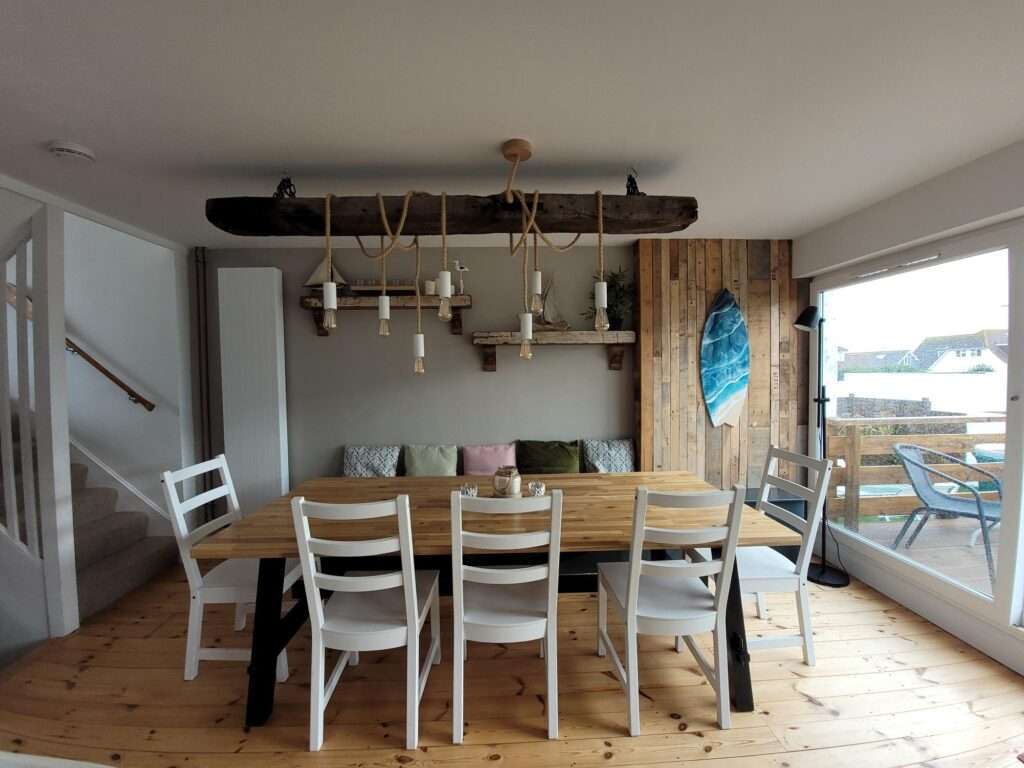 Dining room with wooden table, white chairs, and rustic chandelier. Cushioned bench against the wall, surfboard decor, and large window overlooking a patio with outdoor chairs.