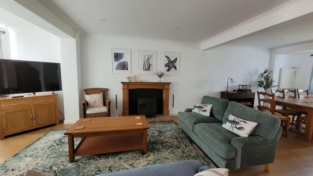 A living room with a green sofa, wooden coffee table, and fireplace. Above the fireplace are four framed artworks. A dining table and chairs are visible in the background.