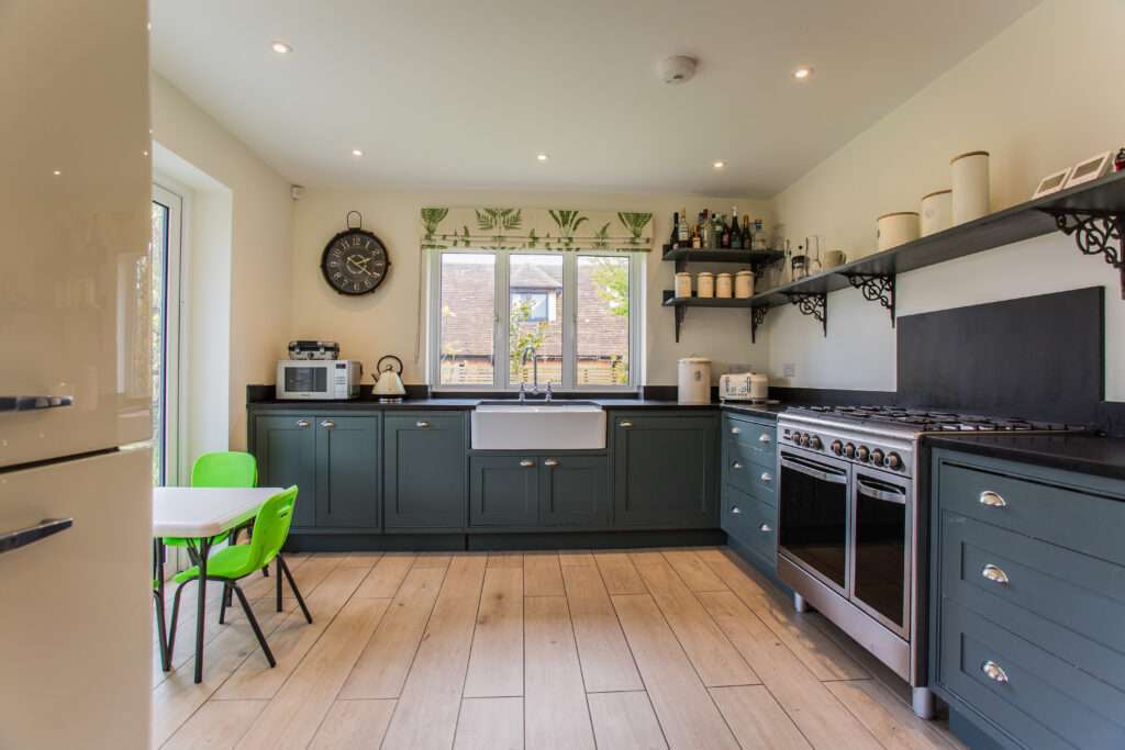 Modern kitchen with green cabinets, large stove, and wooden floor. A small table with bright green chairs is in the corner. Shelves hold jars and bottles, and a clock hangs on the wall.