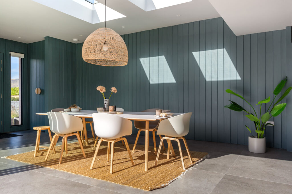 Modern dining room with a long table, white chairs, a woven pendant light, and a large potted plant. Green paneling on the walls, skylights, and natural light create a bright atmosphere.