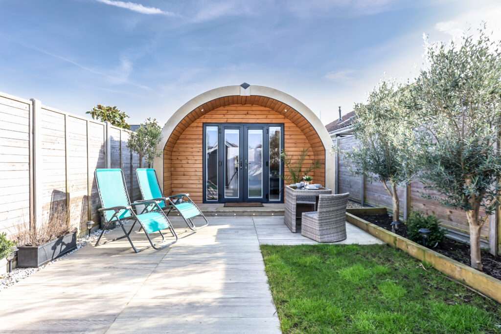 Modern garden room with an arched design, large glass doors, and a patio area featuring two turquoise sun loungers and a round wicker table set, surrounded by a wooden fence and greenery.