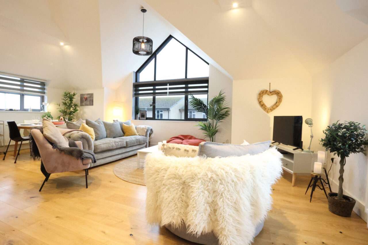 Spacious living room with a large window, modern furniture, gray sofa, beige chairs, TV, and dining area. Decor includes a heart-shaped wall piece and plants. Light wooden floor.