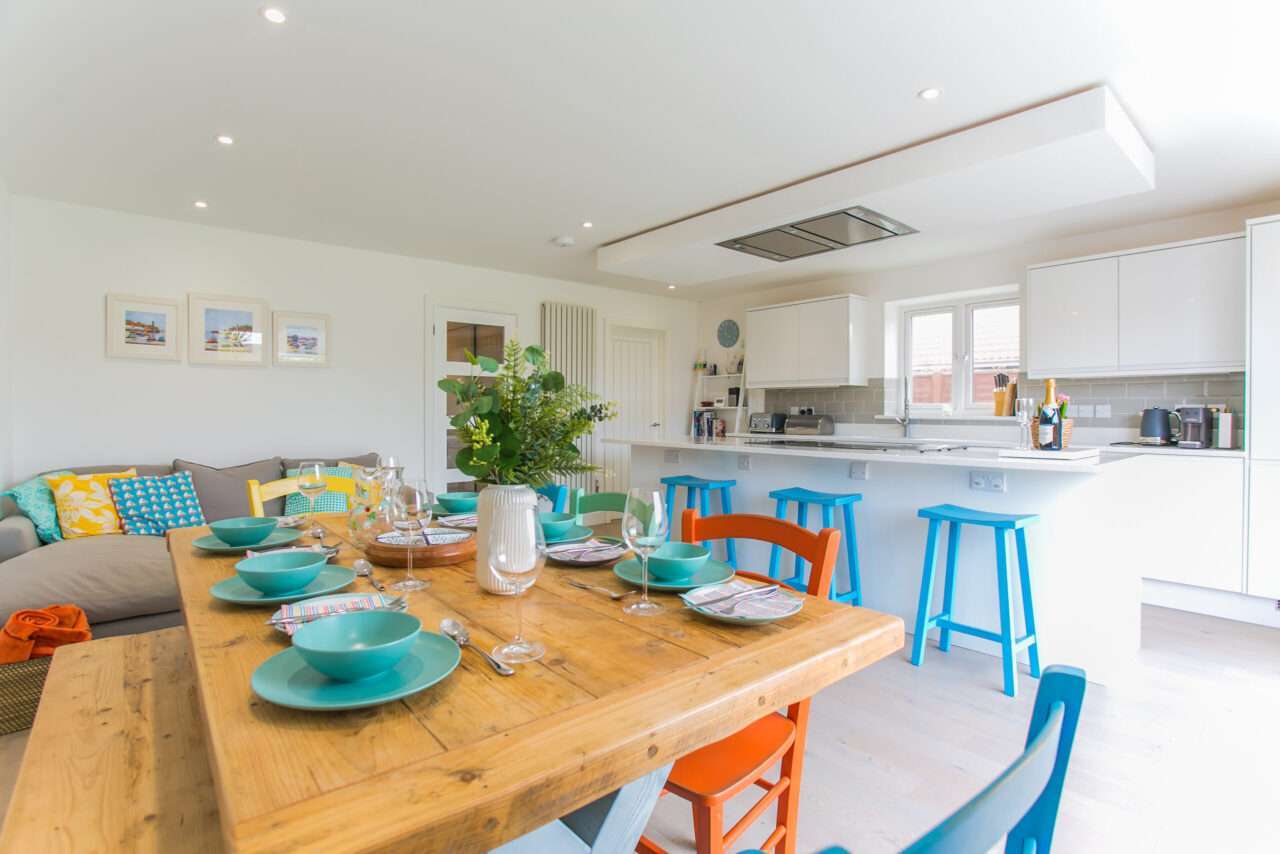 Bright kitchen and dining area with a wooden table set for six. Colorful chairs and stools add contrast. Modern appliances and decor with natural light from the window.