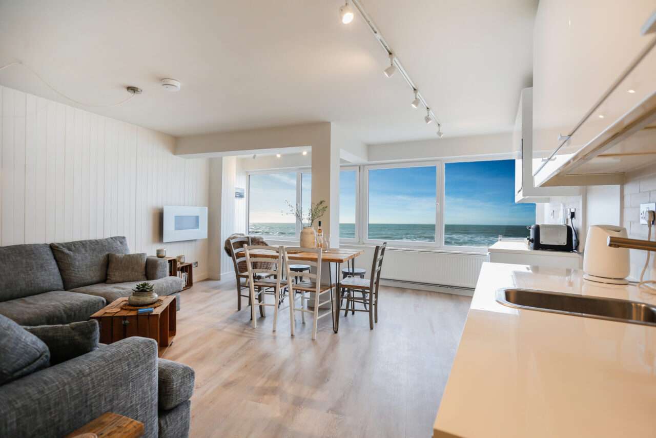 Bright living and dining area with a gray couch, wooden table, and large window offering an ocean view. Modern kitchen with white cabinets and appliances on the right.
