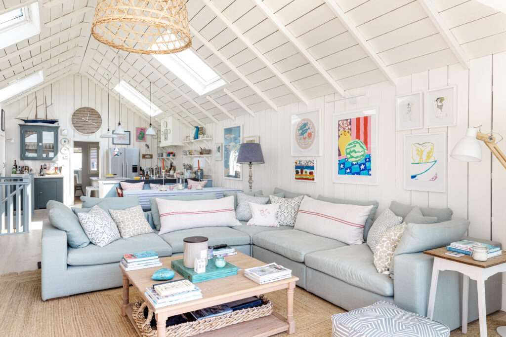 A bright, white living room with vaulted ceilings features a light gray sectional sofa, a wooden coffee table, and eclectic artwork. Skylights and large windows provide ample natural light.