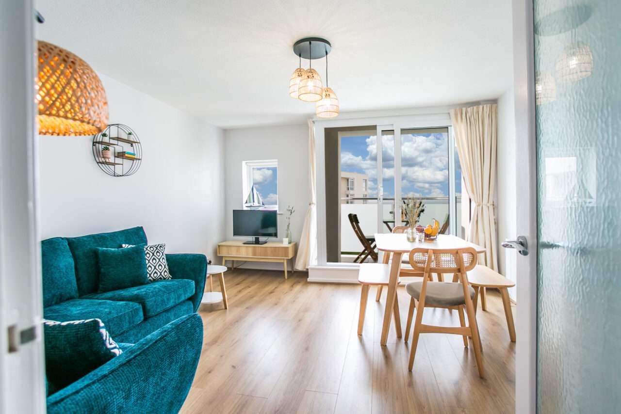 Modern living room with teal sofa, wooden flooring, dining table, and television. Glass door opens to a balcony with a view of clouds. Ceiling lights and decorative items enhance the decor.