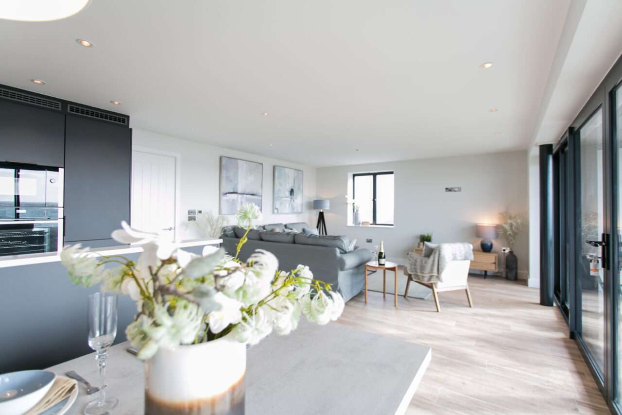 Modern open-plan living area with a kitchen and dining table in the foreground, and a seating area with gray sofas and artwork on the walls in the background. Large windows provide natural light.