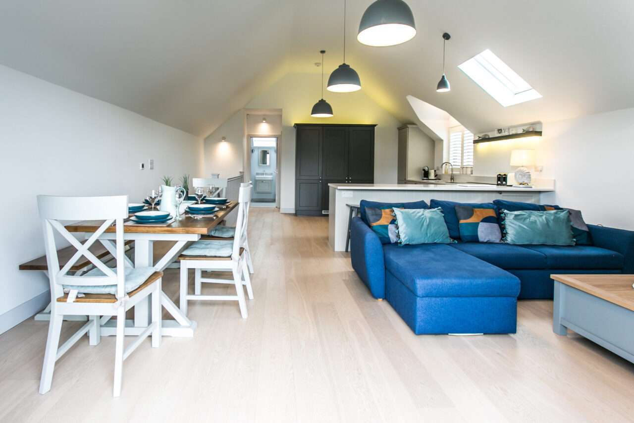 Open-plan living area with a dining table, blue sofa, and kitchen. Pendant lights hang from a sloped ceiling, and natural light enters through large windows.