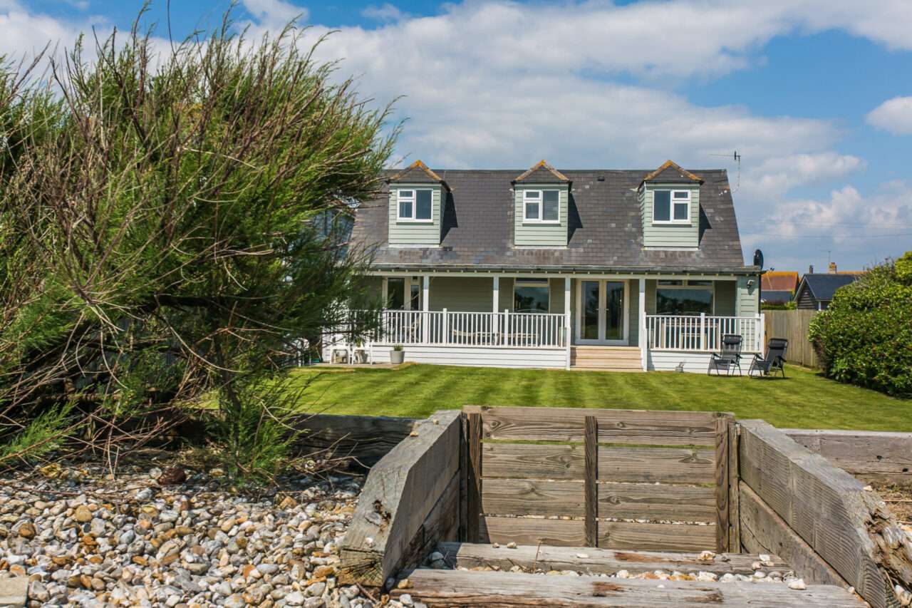 Two-story house with dormer windows and a covered porch, set behind a lawn with shrubs, under a partly cloudy sky. Foreground features a wooden structure and a pebble pathway.