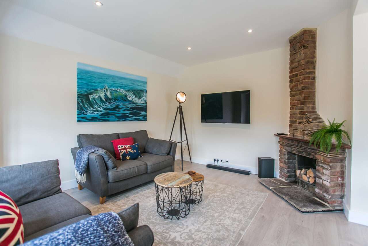 Living room with a brick fireplace, wall-mounted TV, two gray sofas, a round wooden coffee table, and a tripod lamp. A wave painting and Union Jack pillows are also present.