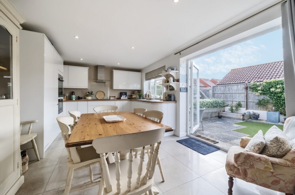 A bright kitchen with a wooden dining table and white chairs, overlooking a patio through open glass doors. The backyard features a small lawn and garden area.